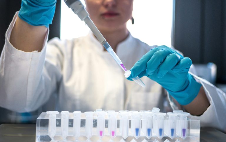 A scientist in a lab coat and teal gloves uses a pipette over a rack of sample vials