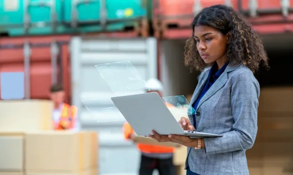 A businesswoman holds a laptop displaying a holographic AR interface in a shipping warehouse