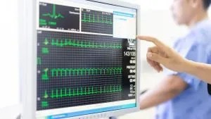 A doctor pointing at a hospital heart-monitor display showing ECG waveforms and vital signs