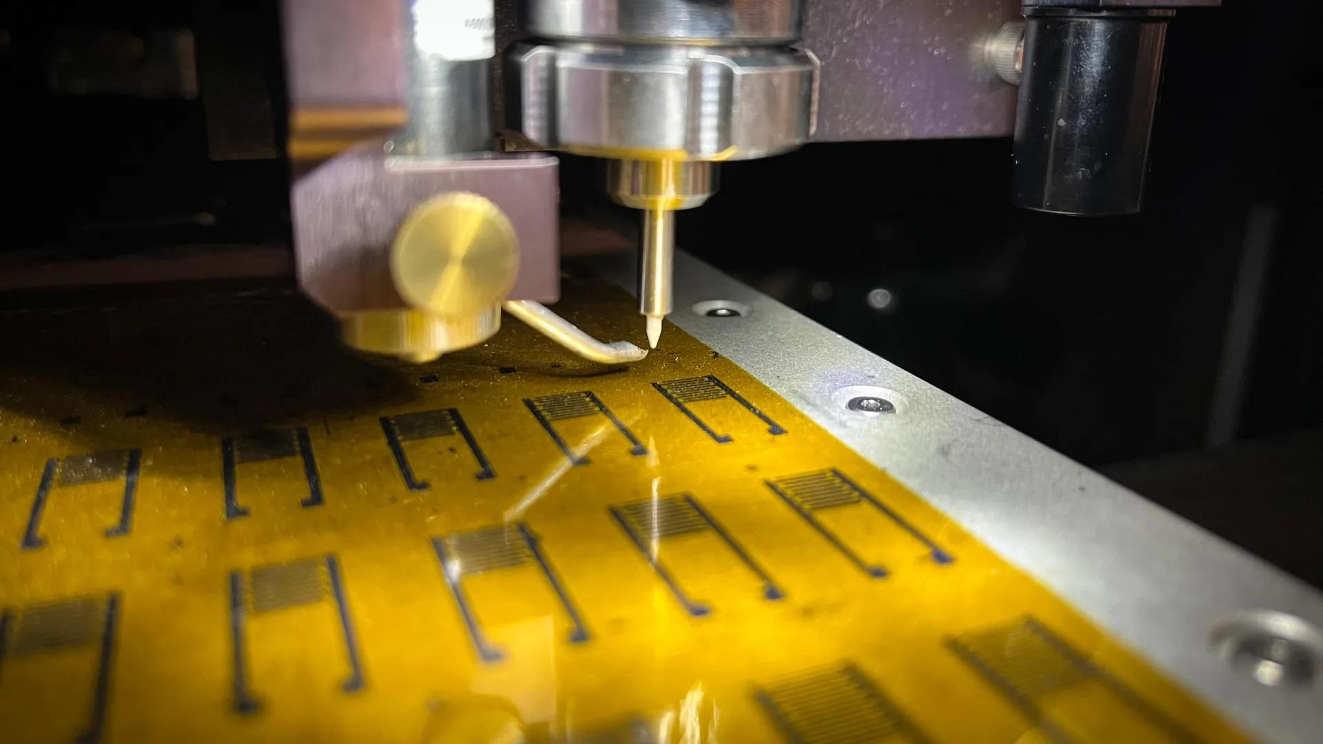 A robotic probe with a gold tip positioned over a flexible yellow circuit sheet printed with rows of comb-like electrode patterns