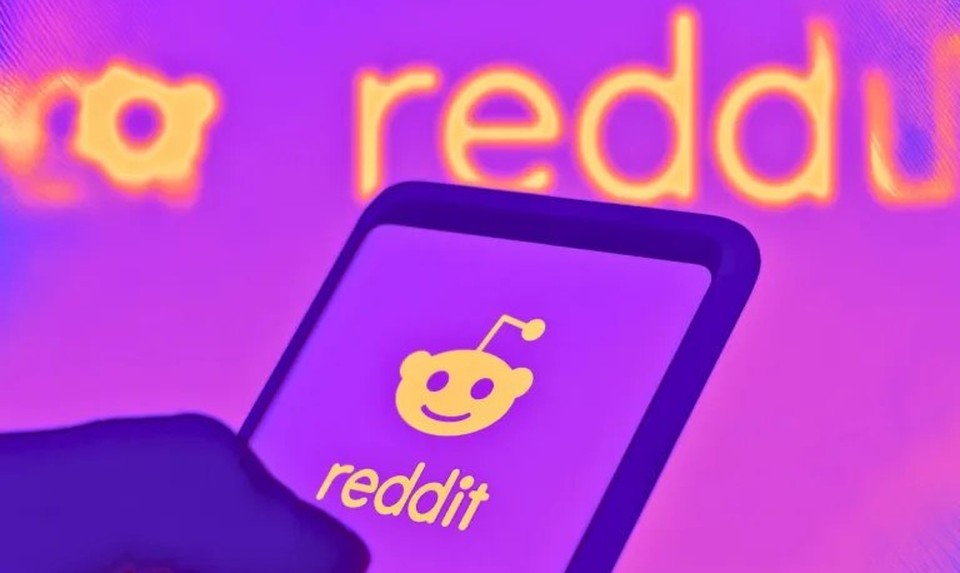 A hand holds a phone showing the Reddit app logo, with the reddit wordmark glowing in the purple-and-orange background