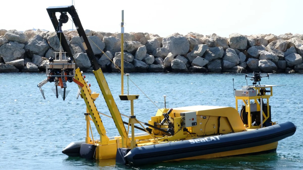 The yellow SeaCAT autonomous surface vessel, fitted with a robotic manipulator arm holding a wire waste basket, floats in a harbor in front of a rock breakwater