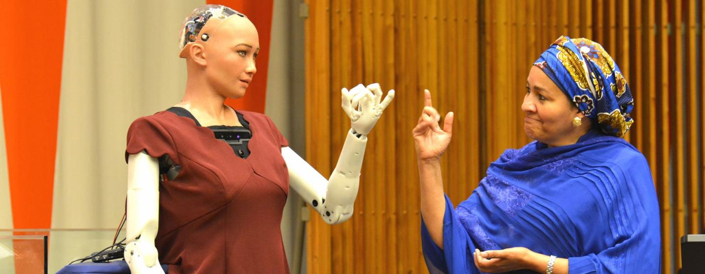 The humanoid robot Sophia, wearing a maroon dress with an exposed mechanical arm, gestures toward a woman in a blue headscarf and robe at a UN event